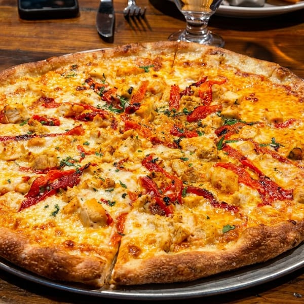A whole chicken and roasted red pepper pizza on a metal pan, on a rustic wooden table.