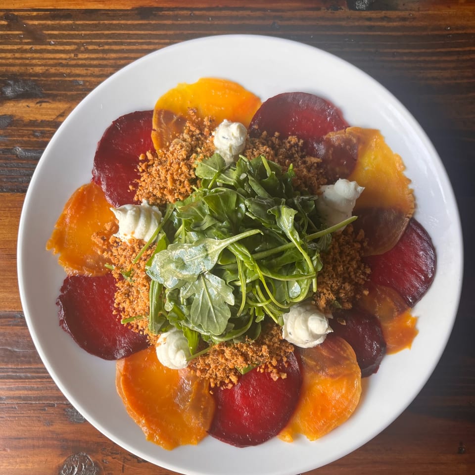 Red and golden beet slices with greens and creamy dollops on a white plate on a wooden table.