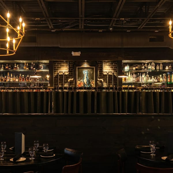 Dark, moody restaurant interior with two large chandeliers, a bar, and set tables.